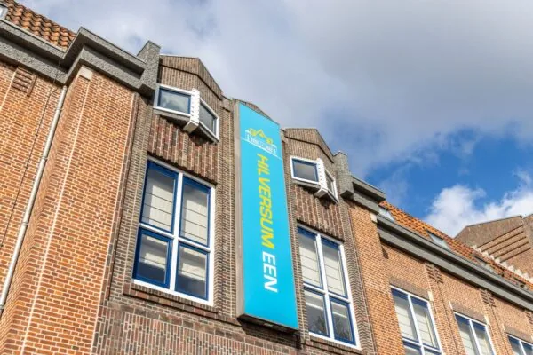 Voorgevel van een bakstenen pand aan de Lage Naarderweg met een blauw gevelbord met de tekst "Hilversum EEN".